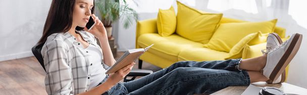 freelancer sitting with legs on table, holding notebook and talking on mobile phone, banner - Photo, Image
