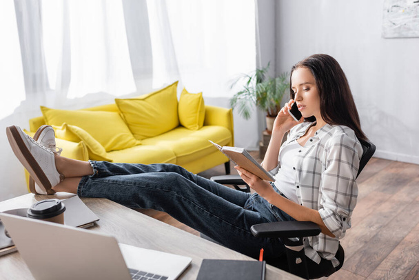 freelancer sitting with legs on table, looking in notebook and talking on smartphone - Photo, image