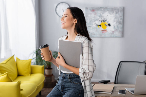 happy freelancer looking away while holding coffee to go and paper folder - Фото, изображение