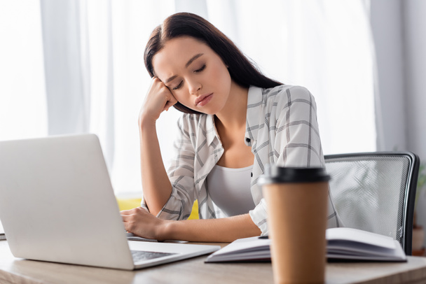exhausted freelancer sitting near laptop and coffee to go on blurred foreground - Photo, Image