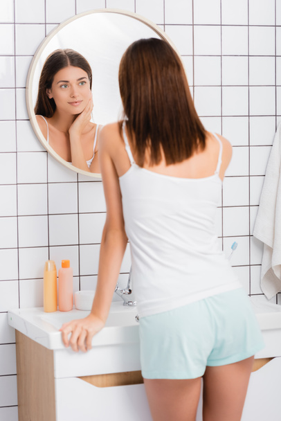 usmívající se žena v bílé kombinéze stojící v koupelně a dívající se do zrcadla - Fotografie, Obrázek