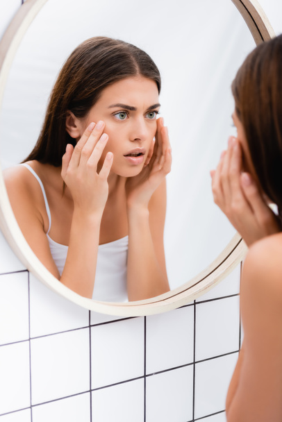 worried young woman looking in mirror and touching face in bathroom, blurred foreground - Foto, Bild