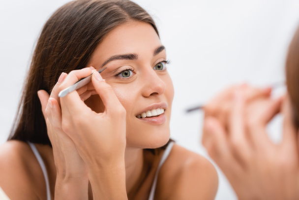 cheerful young woman tweezing eyebrows near mirror, blurred foreground - Photo, Image