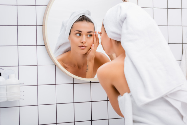 young woman with white towel on head tweezing eyebrows near mirror in bathroom - Foto, imagen