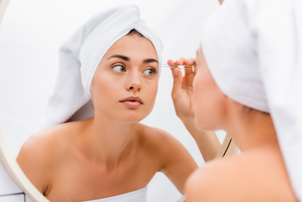 mulher com toalha branca na cabeça tweezing sobrancelhas no banheiro perto do espelho, foreground borrado - Foto, Imagem