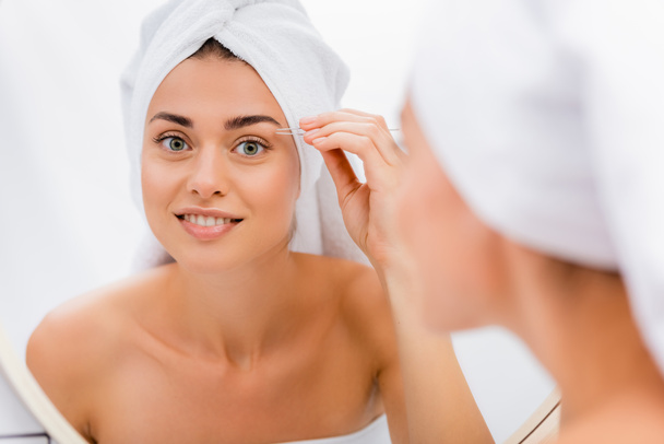 happy woman with white terry towel on head tweezing eyebrows in bathroom, blurred foreground - Photo, Image