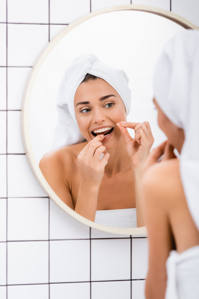 молодая женщина с белым полотенцем на голове зубной нитью рядом с зеркалом в ванной комнате, размытый передний план - Фото, изображение