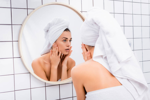 nespokojená mladá žena s bílým ručníkem na hlavě dívá do zrcadla a dotýká tvář v koupelně - Fotografie, Obrázek