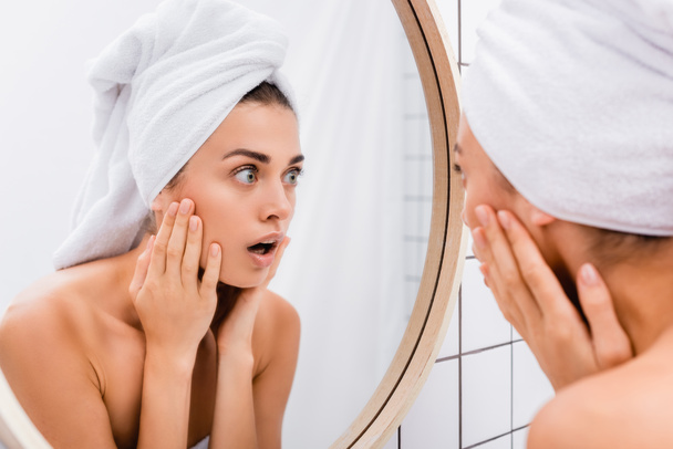shocked young woman with white towel on head touching face while looking in mirror, blurred foreground - Foto, afbeelding