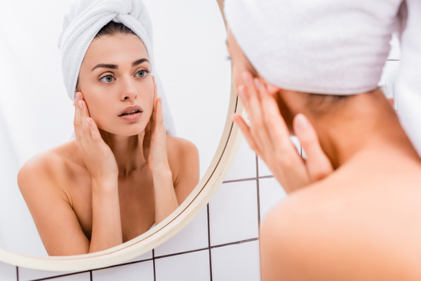 jonge vrouw met handdoek op het hoofd aanraken gezicht tijdens het kijken in spiegel, wazig voorgrond - Foto, afbeelding