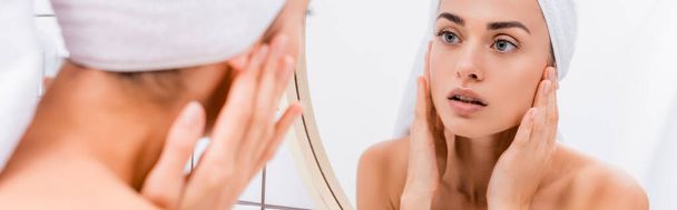 młoda kobieta z białym ręcznikiem na głowie patrząca w lustro i dotykająca twarzy na niewyraźnym pierwszym planie, baner - Zdjęcie, obraz