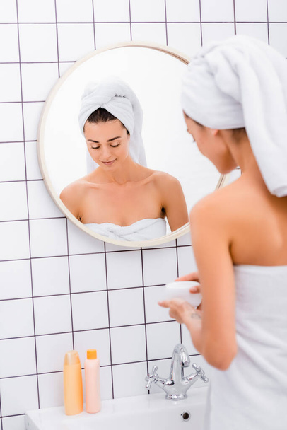 young woman, wrapped in white towels, holding cosmetic cream in bathroom, blurred foreground - Foto, imagen