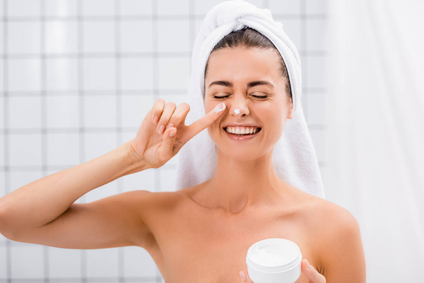 vrolijke vrouw met gesloten ogen aanbrengen gezichtscrème op neus in de badkamer - Foto, afbeelding
