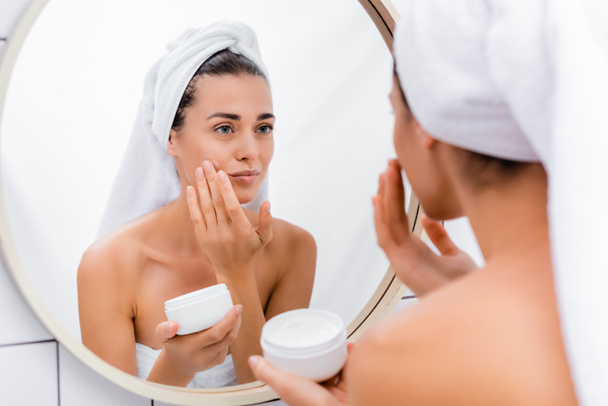 donna con asciugamano bianco sulla testa guardando allo specchio mentre si applica la crema viso in bagno - Foto, immagini