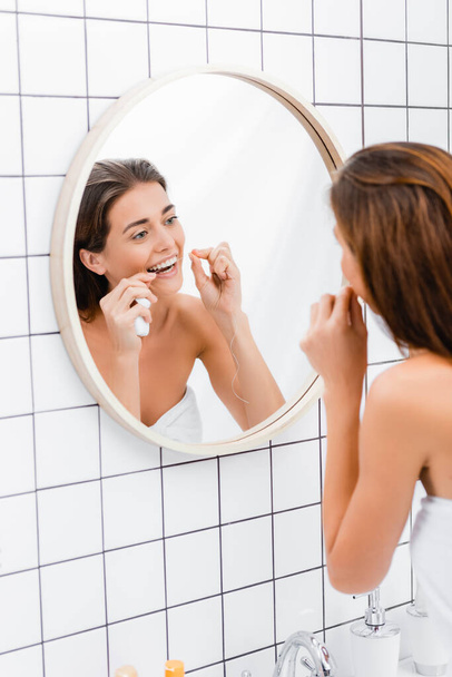 young woman flossing teeth near mirror in bathroom, blurred foreground - Fotó, kép