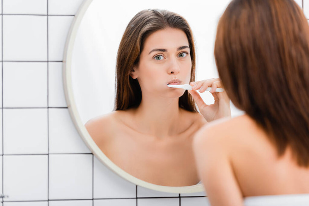 young woman brushing teeth near mirror in bathroom, blurred foreground - Foto, Imagem