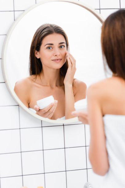 mulher satisfeita aplicando creme facial perto do espelho no banheiro, foreground borrado - Foto, Imagem