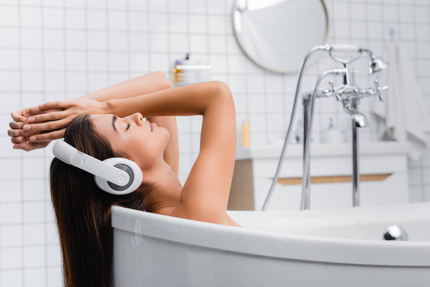 jonge vrouw nemen bad met gesloten ogen en luisteren muziek in draadloze hoofdtelefoon - Foto, afbeelding