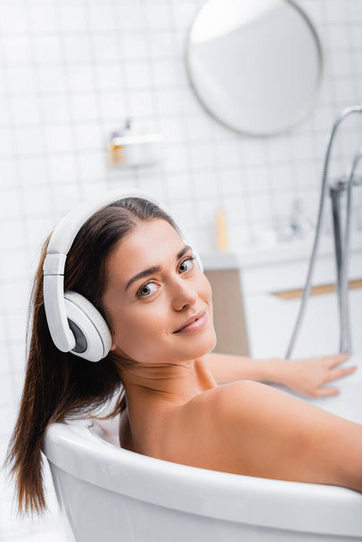 gelukkig jong vrouw kijken naar camera terwijl het nemen van bad en het luisteren van muziek in draadloze hoofdtelefoon - Foto, afbeelding
