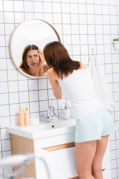 mujer disgustada en pantalones cortos blancos y una sola cara tocando mientras está de pie cerca del espejo en el baño, borrosa primer plano - Foto, imagen