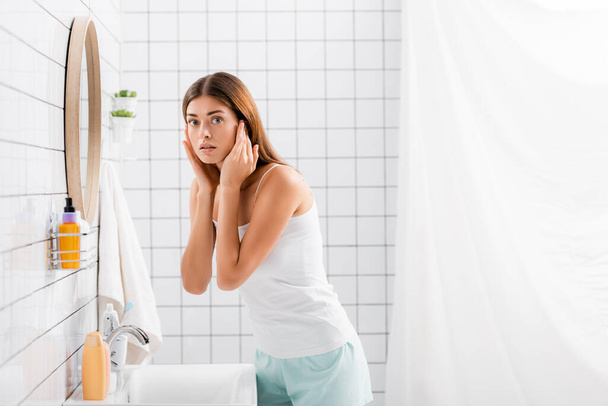 worried young woman in singlet and shorts touching face while looking at camera in bathroom - Foto, afbeelding