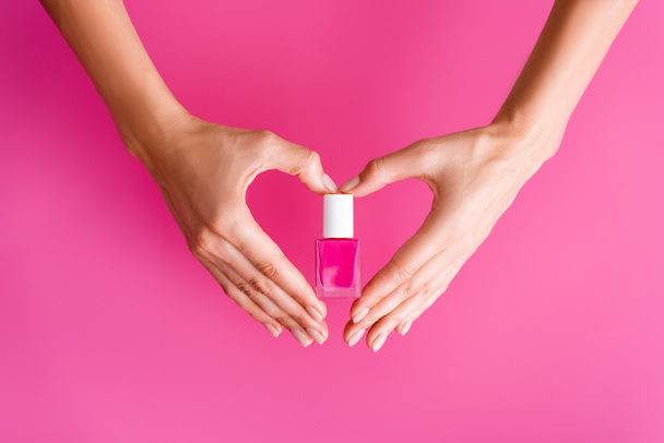 abgeschnittene Ansicht einer Frau, die Herz-Symbol mit Händen macht, während sie eine Flasche Nagellack auf rosa Hintergrund hält - Foto, Bild