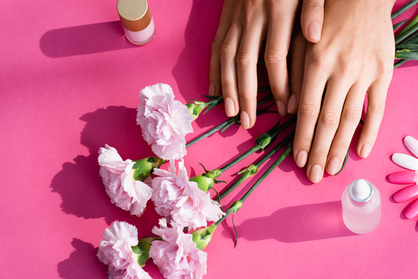 vue du haut des mains féminines près des fleurs d'oeillets, palette de faux ongles, flacons avec décapant pour cuticules et vernis à ongles sur fond rose - Photo, image