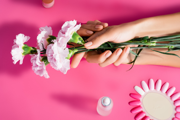 cropped view of woman holding carnation flowers near palette of artificial nails and cuticle remover on pink background - Photo, Image