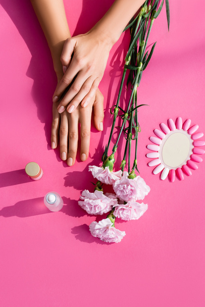 vista superior das mãos femininas perto de flores do cravo, paleta de unhas artificiais, removedor de cutículas e esmalte no fundo rosa - Foto, Imagem