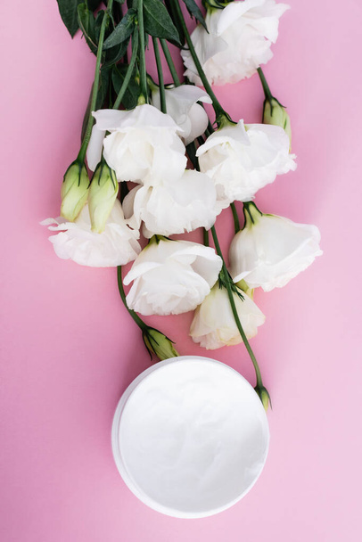 Draufsicht auf kosmetische Creme und weiße Eustoma-Blüten auf rosa Hintergrund - Foto, Bild
