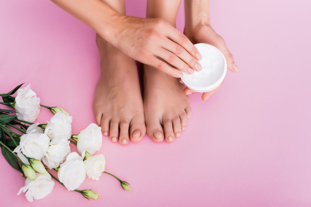 cropped view of barefoot woman holding cosmetic cream near white eustoma flowers on pink background - Fotografie, Obrázek