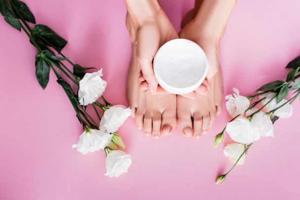 top view of female hands with cosmetic cream near feet and white eustoma flowers on pink background - 写真・画像