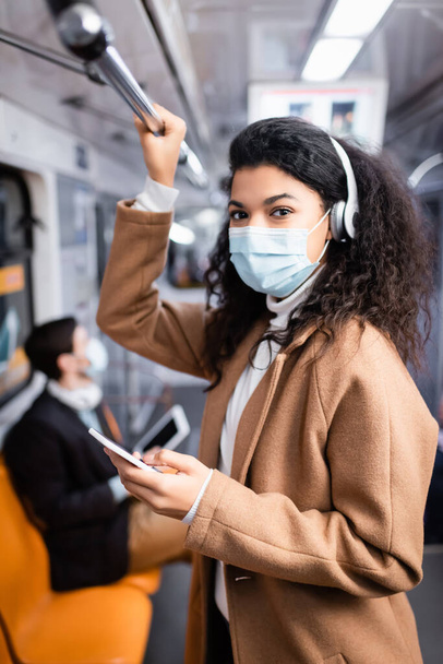 kudrnaté africké americká žena v lékařské masce poslech hudby a pomocí smartphone v metru  - Fotografie, Obrázek