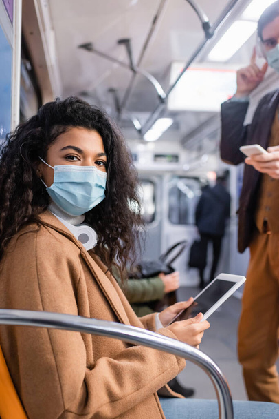 afro-américaine en masque médical à l'aide d'une tablette numérique près de l'homme dans le métro avec fond flou  - Photo, image