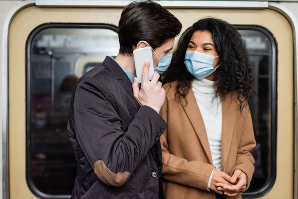Afrikanerin mit medizinischer Maske sieht Freund in U-Bahn beim Smartphone-Gespräch an - Foto, Bild