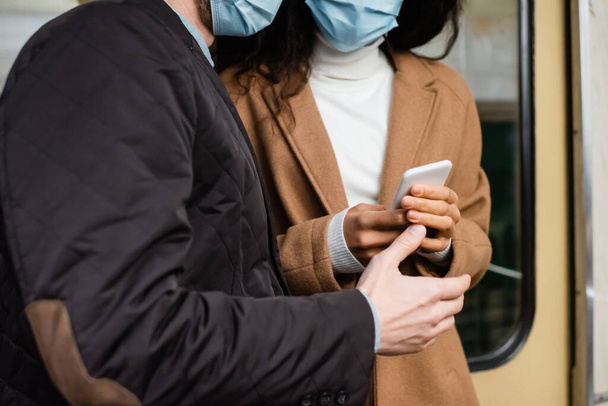 обрезанный вид на африканскую американку в медицинской маске с помощью смартфона рядом с мужчиной в метро  - Фото, изображение