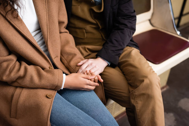 abgeschnittene Ansicht eines gemischtrassigen Paares, das Händchen hält, während es im Waggon der U-Bahn sitzt - Foto, Bild