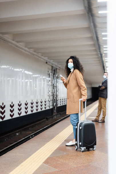comprimento total da mulher americana africana em máscara médica de pé com bagagem perto do homem no metrô  - Foto, Imagem