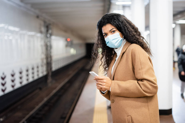 σγουρά αφροαμερικανή γυναίκα με ιατρική μάσκα κρατώντας smartphone στο μετρό - Φωτογραφία, εικόνα