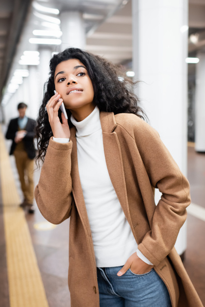 mujer afroamericana mordiendo los labios mientras habla en el teléfono inteligente y mirando hacia otro lado en el metro en un fondo borroso  - Foto, imagen