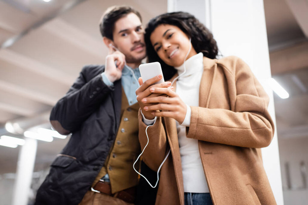 vista en ángulo bajo de pareja multicultural con smartphone escuchando música en auriculares en metro sobre fondo borroso - Foto, imagen