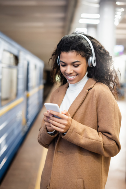 улыбающаяся африканская американка в беспроводных наушниках с помощью смартфона во время прослушивания музыки в метро  - Фото, изображение