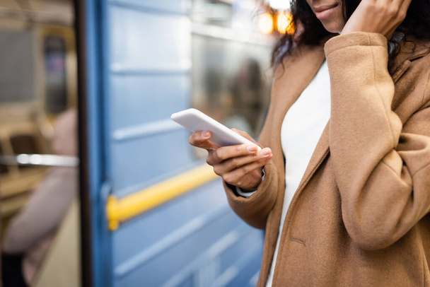 vista recortada de mujer afroamericana usando teléfono inteligente cerca de vagón en metro sobre fondo borroso  - Foto, imagen