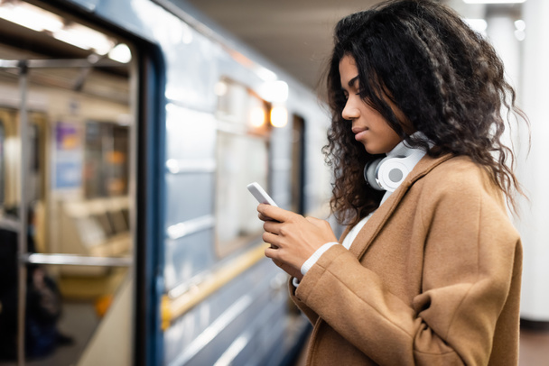 zijaanzicht van Afrikaanse Amerikaanse vrouw in draadloze hoofdtelefoon met behulp van smartphone in de metro  - Foto, afbeelding