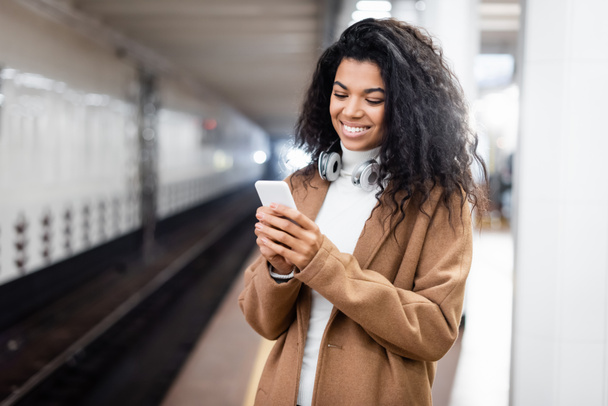smiling african american woman in wireless headphones using smartphone in subway  - Photo, Image
