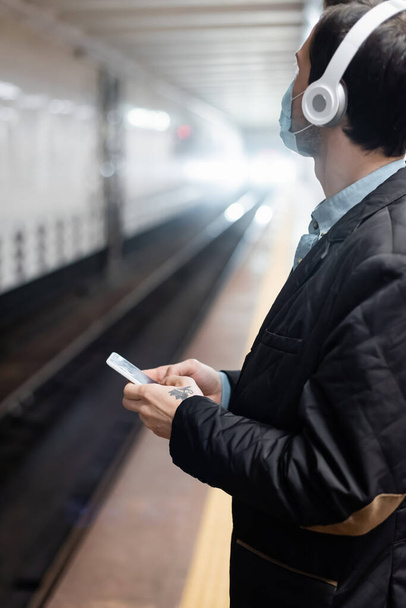 wytatuowany mężczyzna w masce medycznej i słuchawkach trzymających smartfona w metrze  - Zdjęcie, obraz