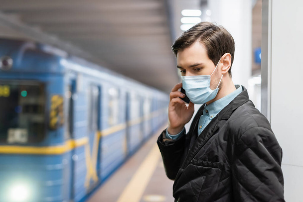 mężczyzna w masce medycznej mówi na smartfonie w pobliżu wagonu metra na rozmytym tle - Zdjęcie, obraz