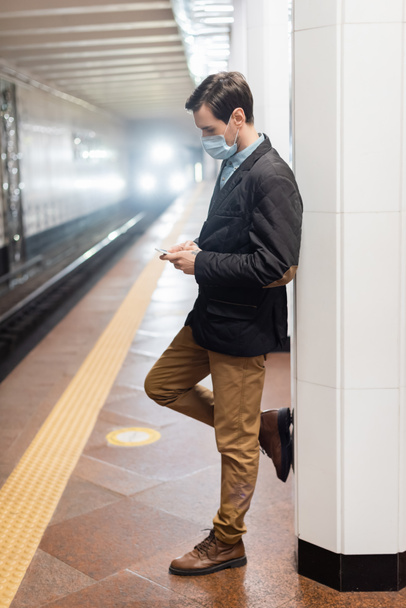 Mann in medizinischer Maske steht und Smartphone in U-Bahn-Halle hält - Foto, Bild