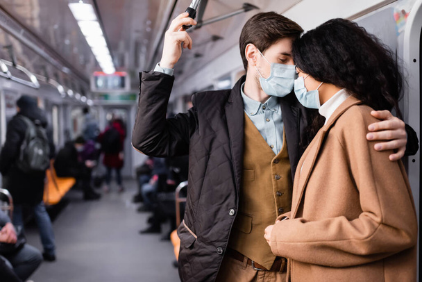 man in medical mask hugging african american girlfriend in wagon of metro with people on blurred background - Photo, image