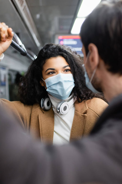 Αφροαμερικανή γυναίκα και άντρας με ιατρικές μάσκες κοιτάζονται στο βαγόνι του μετρό. - Φωτογραφία, εικόνα